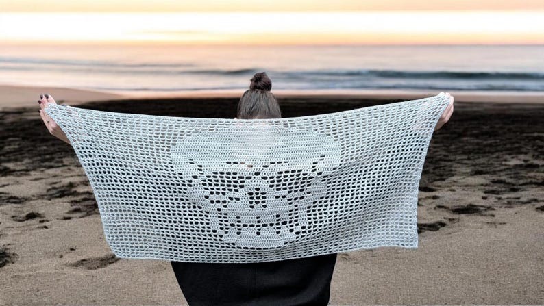 May include: A light blue, open-weave crochet shawl with a skull design. The shawl is held up against a beach backdrop, with the ocean and sky visible in the background. The crochet pattern is visible.