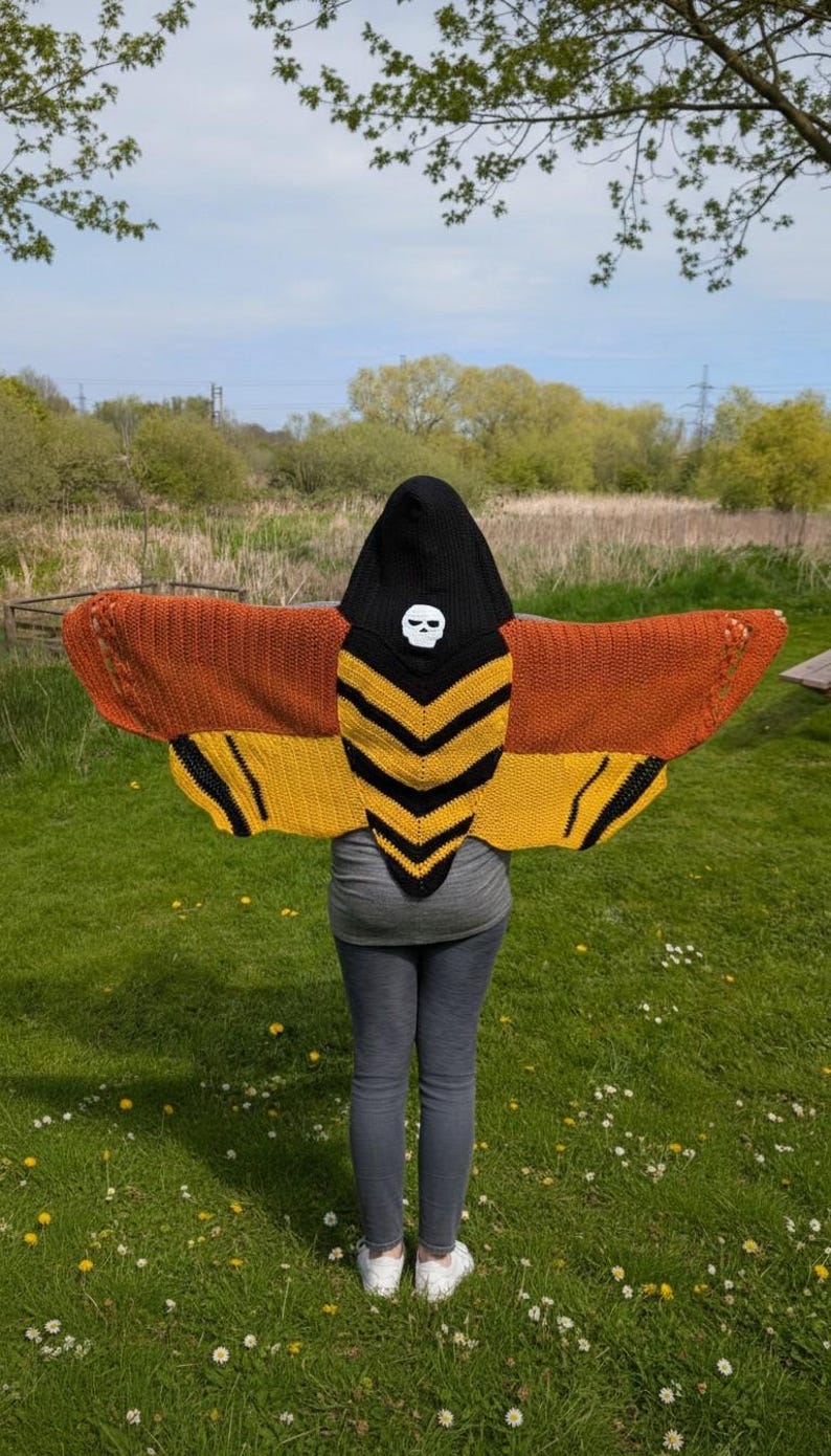 May include: A hooded shawl resembling a moth, with brown, yellow, and black wings. The hood is black with a white skull design. The shawl is worn outdoors, with a green grassy background and a blue sky.