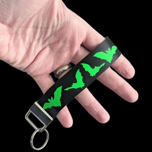 May include: Black wristlet keychain with a repeating pattern of neon green bat silhouettes. The keychain has a silver-colored metal clasp and key ring. The wrist strap is made of a textured material.