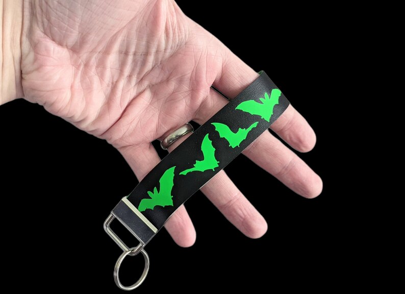 May include: Black wristlet keychain with a repeating pattern of neon green bat silhouettes. The keychain has a silver-colored metal clasp and key ring. The wrist strap is made of a textured material.