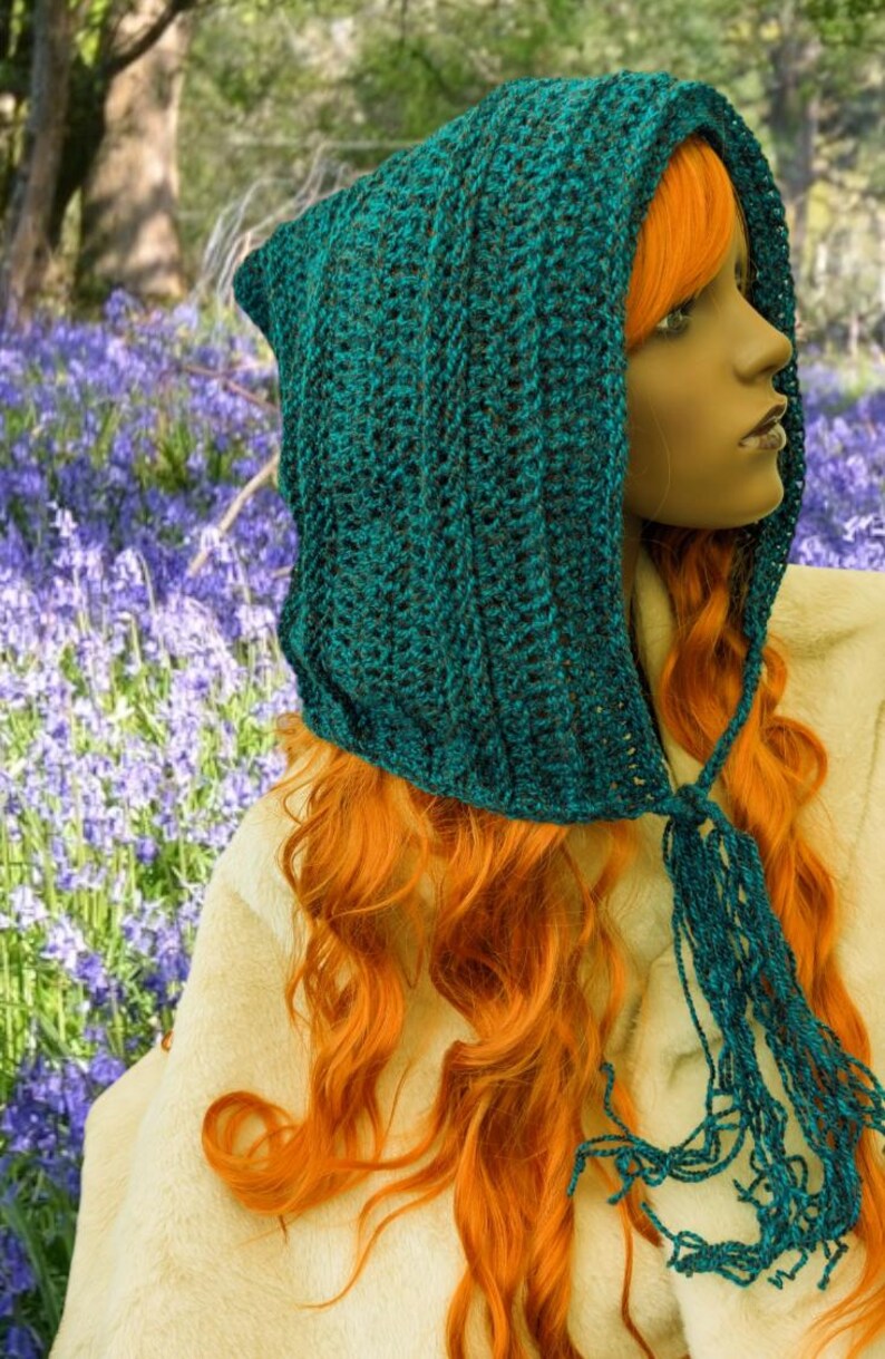 May include: A teal blue crocheted hooded scarf with a long fringe. The scarf is worn by a person with long, bright orange hair.