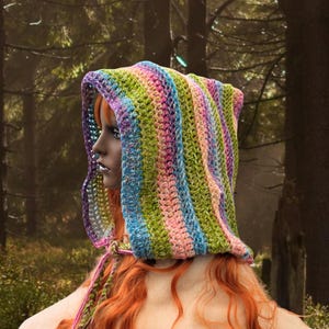 May include: A colourful striped crochet hooded scarf in shades of green, pink, blue, and purple. The scarf is worn by a person with long red hair.