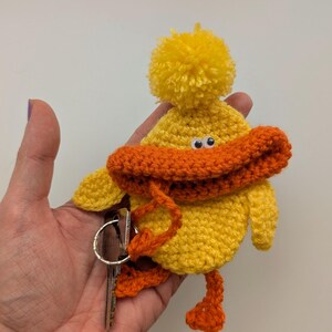 May include: A yellow crocheted duck keychain with an orange beak and feet, and a yellow pom-pom on its head. The keychain is attached to a silver key ring with keys.