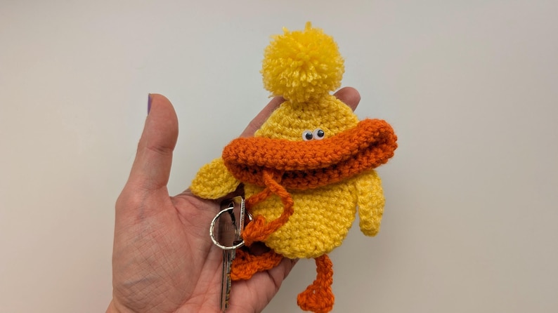 May include: A yellow crocheted duck keychain with an orange beak and feet, and a yellow pom-pom on its head. The keychain is attached to a silver key ring with keys.