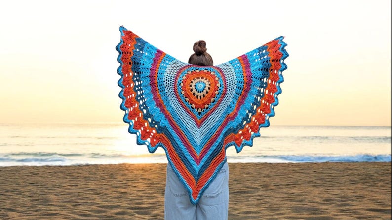 May include: A colorful, handmade crochet shawl with a vibrant design. The shawl features a central circular pattern in orange and blue, surrounded by concentric rings of blue, orange, and pink. The shawl is displayed against a beach backdrop.