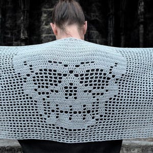 May include: A light gray, open-weave crochet shawl with a butterfly design. The shawl is held up, showcasing the intricate pattern. The background is a dark, stone structure.