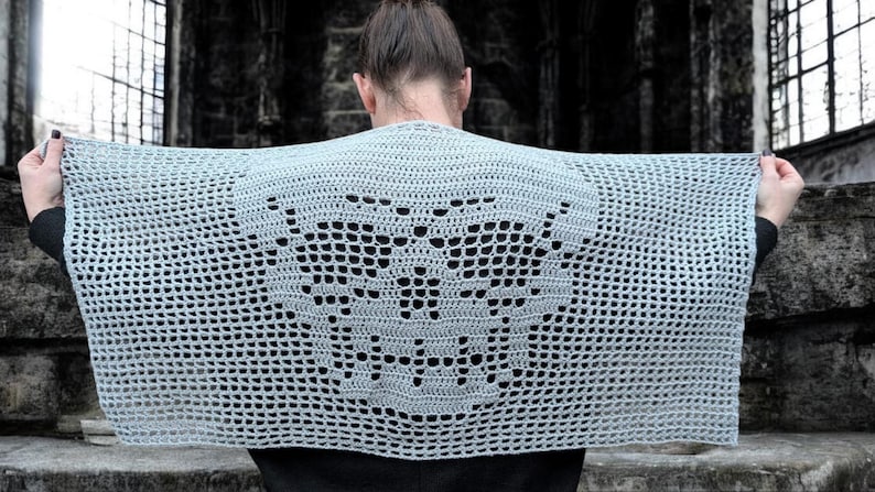 May include: A light gray, open-weave crochet shawl with a butterfly design. The shawl is held up, showcasing the intricate pattern. The background is a dark, stone structure.