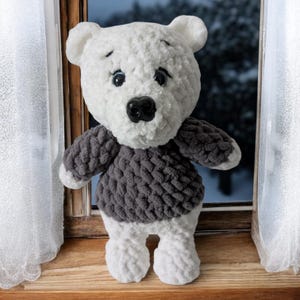 May include: A handmade white and gray teddy bear stands on a wooden windowsill. The bear has a white head and legs, a gray sweater, and black eyes and nose. The background shows a snowy scene outside a window.