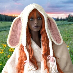 May include: A white crocheted bunny hat with pink ears and long white tassels. The hat is worn by a mannequin with long red hair.