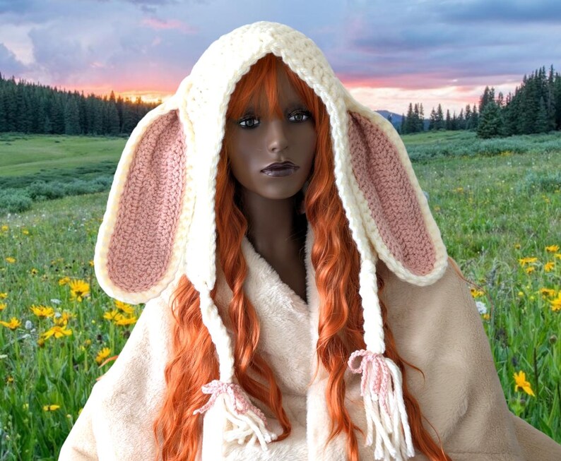 May include: A white crocheted bunny hat with pink ears and long white tassels. The hat is worn by a mannequin with long red hair.