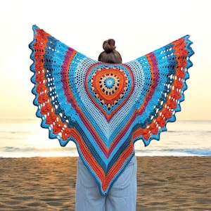 May include: A colorful, handmade crochet shawl in shades of blue, orange, and pink. The shawl features a heart-shaped design in the center and a scalloped edge. The person wearing the shawl is standing on a beach with the ocean in the background.