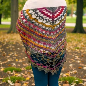 May include: A multi-coloured crocheted shawl in shades of grey, pink, yellow, and green. The openwork design has a scalloped edge and is draped over the shoulders. The shawl is triangular and appears to be handmade.