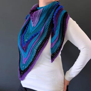 May include: A handmade crochet shawl in shades of teal, purple, and black. The triangular shawl features a striped pattern with textured details and a scalloped edge. The shawl is worn over a white long-sleeved shirt.