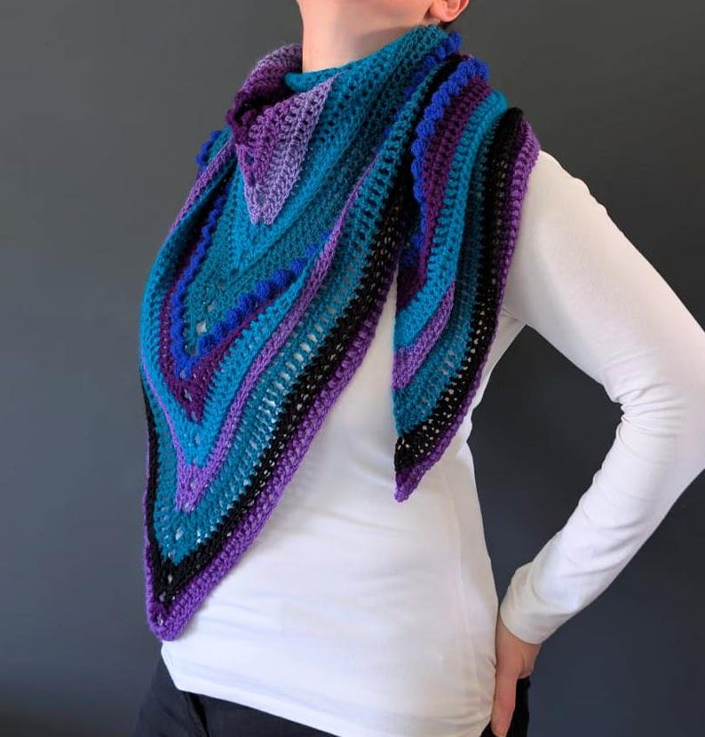May include: A handmade crochet shawl in shades of teal, purple, and black. The triangular shawl features a striped pattern with textured details and a scalloped edge. The shawl is worn over a white long-sleeved shirt.