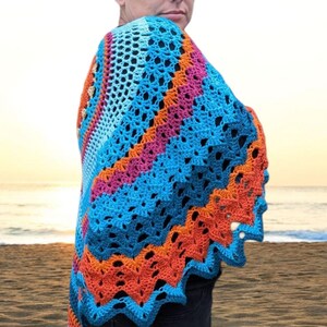 May include: A colorful crochet shawl in shades of blue, orange, and pink. The shawl has a scalloped edge and a geometric pattern. The person is standing on a beach with the ocean and sky in the background.