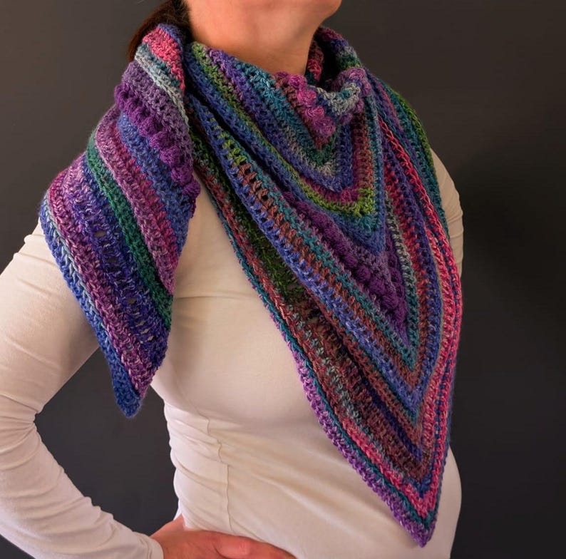 Pu&ograve; includere: Uno scialle all'uncinetto fatto a mano e colorato, nei toni del blu, viola, verde e rosa. Lo scialle triangolare &egrave; drappeggiato intorno al collo, mostrando un motivo a punto strutturato. Il filato sembra essere una miscela di pi&ugrave; colori, creando un effetto a righe.