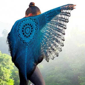 May include: A handmade, blue and black crochet shawl with a circular design on the back. The shawl is draped over the shoulders, with the edges spread out. The background is a blurred view of a forest.