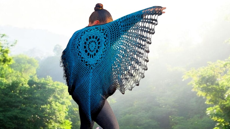 May include: A handmade, blue and black crochet shawl with a circular design on the back. The shawl is draped over the shoulders, with the edges spread out. The background is a blurred view of a forest.