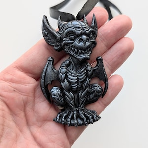 May include: A dark gray gargoyle pendant with a black ribbon. The pendant features a detailed design of a seated gargoyle with large wings, horns, and a toothy grin. The gargoyle is intricately sculpted with a textured finish.