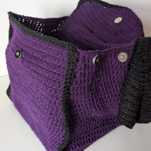 May include: A handmade crochet bag in a deep purple color with black trim. The bag features a textured stitch pattern and is secured with silver magnetic snap closures. The bag is open, revealing the interior.