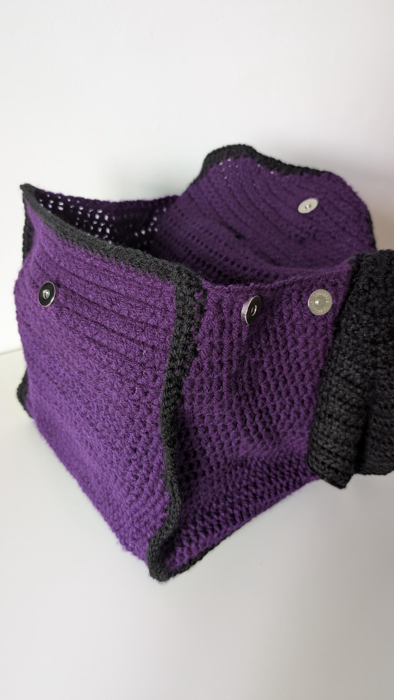 May include: A handmade crochet bag in a deep purple color with black trim. The bag features a textured stitch pattern and is secured with silver magnetic snap closures. The bag is open, revealing the interior.