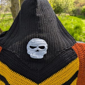 May include: A black crochet hooded cowl with a gray skull applique. The cowl features yellow and black chevron stripes and is paired with an orange crochet shawl. The background is a natural outdoor setting.