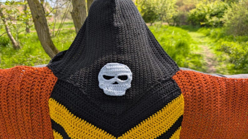 May include: A black crochet hooded cowl with a gray skull applique. The cowl features yellow and black chevron stripes and is paired with an orange crochet shawl. The background is a natural outdoor setting.