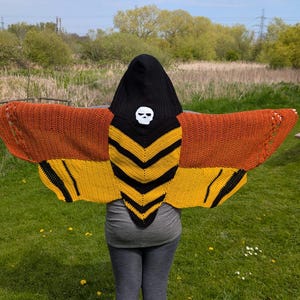 May include: A knitted hooded shawl in the shape of a moth. The shawl features orange and yellow wings with black accents. The hood is black with a white skull. The person is standing in a grassy field.