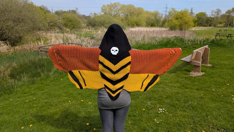 May include: A knitted hooded shawl in the shape of a moth. The shawl features orange and yellow wings with black accents. The hood is black with a white skull. The person is standing in a grassy field.