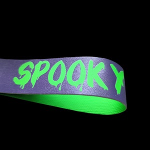 May include: A purple band with the word "SPOOKY" in dripping neon green letters. The underside of the band is bright green. The background is black.