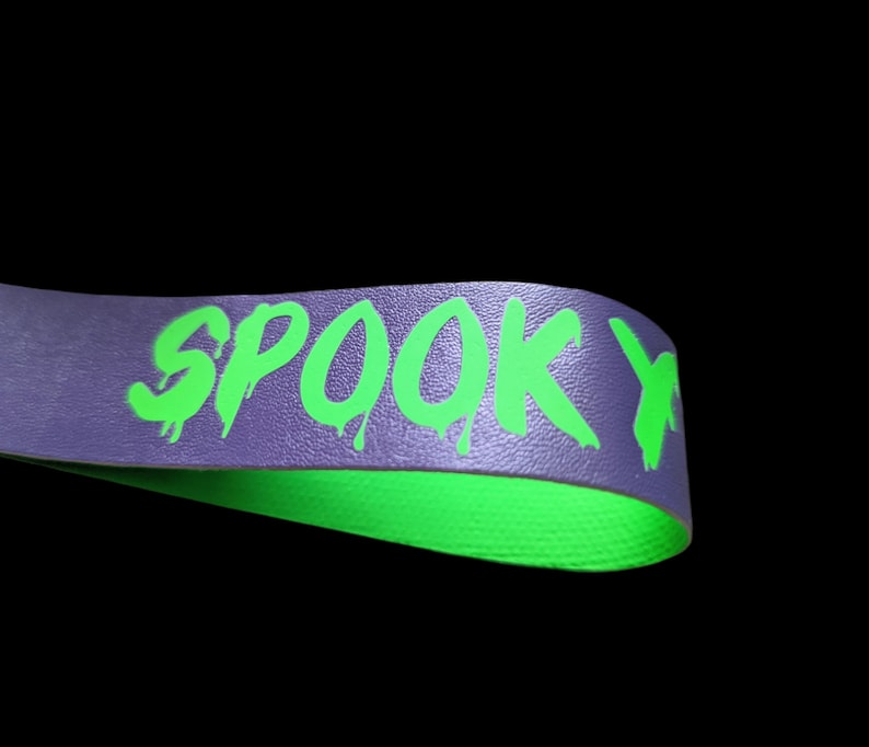 May include: A purple band with the word "SPOOKY" in dripping neon green letters. The underside of the band is bright green. The background is black.