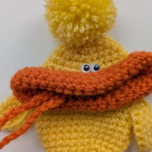 May include: A handmade yellow crochet duck with an orange beak and a yellow pom-pom on top. The duck has googly eyes and orange yarn strings. This whimsical item is perfect for a child's toy or nursery decor.