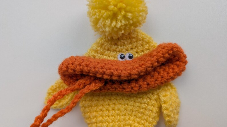 May include: A handmade yellow crochet duck with an orange beak and a yellow pom-pom on top. The duck has googly eyes and orange yarn strings. This whimsical item is perfect for a child's toy or nursery decor.