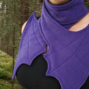 May include: Purple fleece scarf with a bat wing design. The scarf is folded and draped around a person's neck.