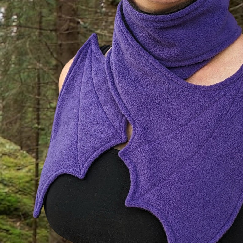 May include: Purple fleece scarf with a bat wing design. The scarf is folded and draped around a person's neck.