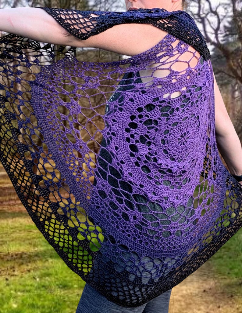 May include: A handmade crochet shawl in a gradient of purple to black. The openwork design features a spiral pattern and a delicate, lacy texture. The shawl is draped over the shoulders, showcasing the intricate craftsmanship and colour transition.