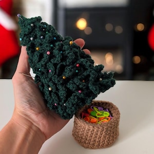 Puede incluir: Un árbol de Navidad de ganchillo verde oscuro decorado con cuentas y alfileres de colores. Debajo hay una pequeña cesta de ganchillo marrón llena de marcadores de punto de colores. El árbol mide unos 15 cm de altura.