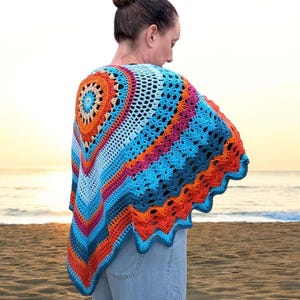 Op de afbeelding: Een kleurrijke, gehaakte sjaal in tinten blauw, oranje en roze. De sjaal heeft een cirkelvormig ontwerp op de rug en een geschulpte rand. De foto is genomen op een strand bij zonsondergang.