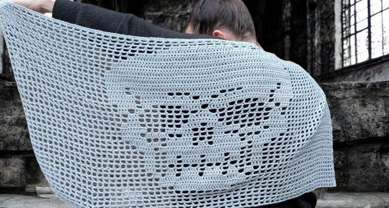 May include: A light gray crocheted shawl with a skull design. The open-weave pattern creates a delicate, airy texture. The shawl is draped over the shoulders, showcasing the intricate skull motif. The background is a blurred outdoor setting.