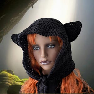 Puede incluir: Un gorro negro de ganchillo con capucha y orejas de gato. La capucha est&aacute; atada al cuello con un cord&oacute;n negro. El gorro lo lleva puesto un maniqu&iacute; con el pelo largo y rojo.