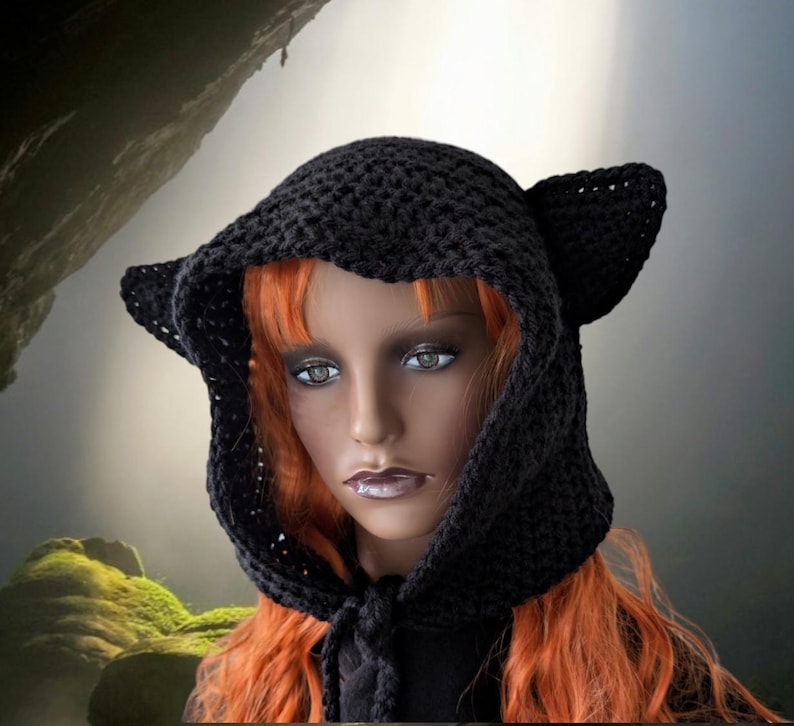 Puede incluir: Un gorro negro de ganchillo con capucha y orejas de gato. La capucha est&aacute; atada al cuello con un cord&oacute;n negro. El gorro lo lleva puesto un maniqu&iacute; con el pelo largo y rojo.