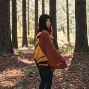 May include: A hooded, open-front sweater in shades of black, yellow, and rust. The sweater has a striped pattern on the sleeves and a solid black hood. The person is wearing black pants and brown boots, standing in a forest setting.