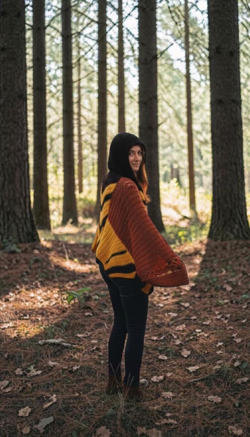 May include: A hooded, open-front sweater in shades of black, yellow, and rust. The sweater has a striped pattern on the sleeves and a solid black hood. The person is wearing black pants and brown boots, standing in a forest setting.