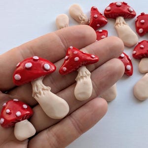 May include: A selection of miniature mushroom ornaments. The mushrooms feature red caps with white spots and beige stems. These small decorative items are likely made from clay or a similar material.