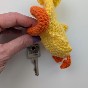 May include: A handmade yellow and orange crochet duck keychain. The duck has orange feet and a beak, with a key attached. The keychain is made of yarn and is a cute, whimsical accessory.