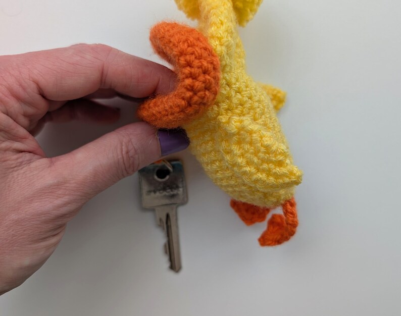 May include: A handmade yellow and orange crochet duck keychain. The duck has orange feet and a beak, with a key attached. The keychain is made of yarn and is a cute, whimsical accessory.
