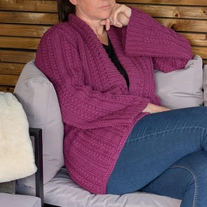 May include: A magenta-colored, open-front cardigan with bell sleeves. The knitted sweater features a textured pattern and is paired with blue jeans. The person is seated on a gray sofa with a white pillow.