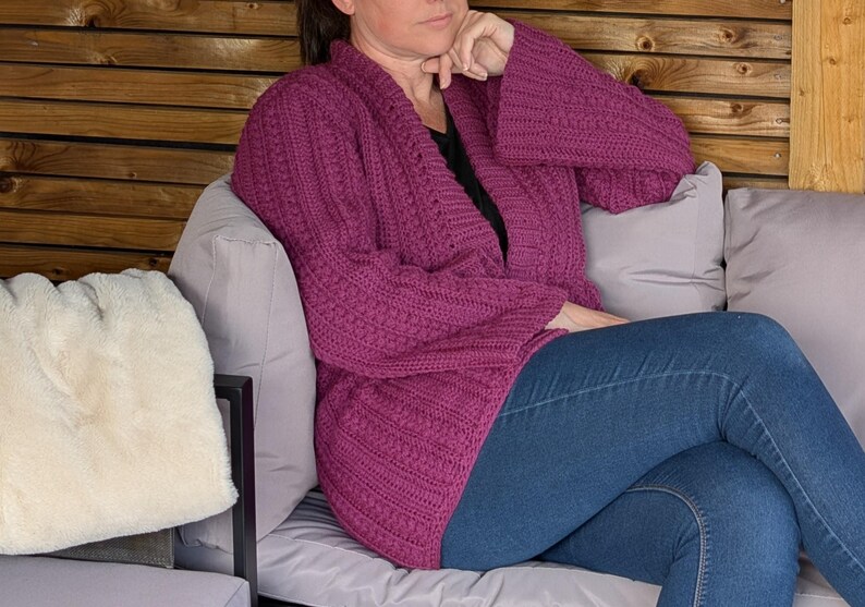 May include: A magenta-colored, open-front cardigan with bell sleeves. The knitted sweater features a textured pattern and is paired with blue jeans. The person is seated on a gray sofa with a white pillow.