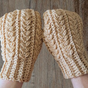 May include: A pair of light tan crochet mittens. The mittens have a textured, woven pattern and a ribbed cuff. The mittens are displayed on a wooden surface.