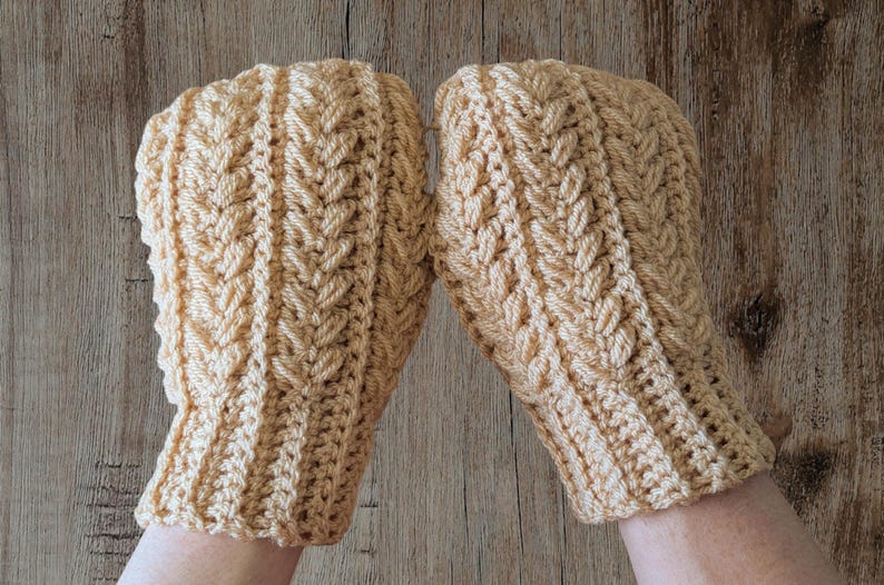 May include: A pair of light tan crochet mittens. The mittens have a textured, woven pattern and a ribbed cuff. The mittens are displayed on a wooden surface.